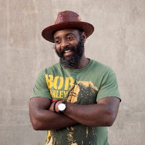 Reginald Dwayne Betts' headshot. He is wearing a brown hat and a Bob Marley shirt. His arms are crossed and he is smiling.